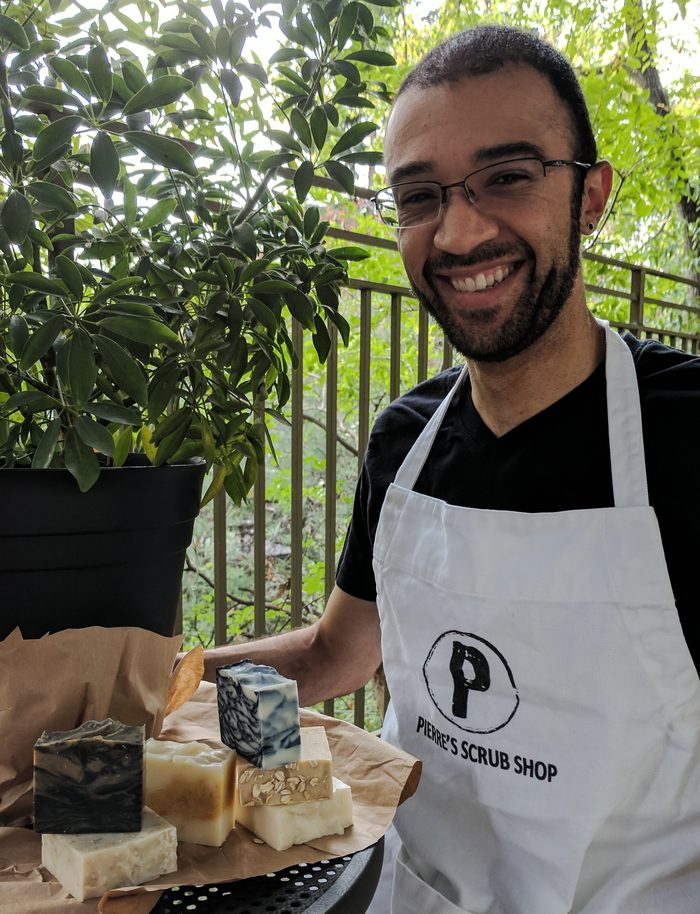 Roberto Pierre Roque standing behind a market stall in a Pierre's Scrub Shop apron with bars of soap laid out on a board