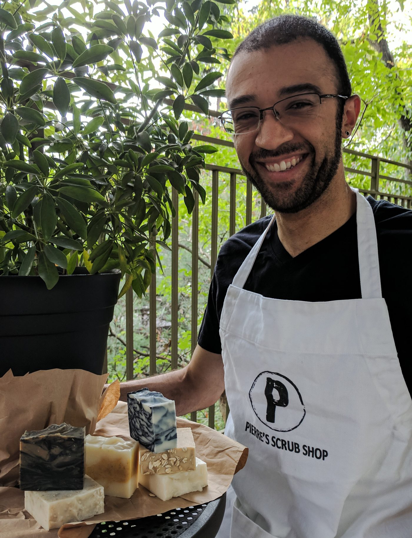 Roberto Pierre Roque standing behind a market stall in a Pierre's Scrub Shop apron with bars of soap laid out on a board
